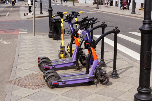 Eine Reihe von E-Scootern, die auf dem Bürgersteig geparkt sind, mit Fußgängern, Radfahrern, einem Müllcontainer im Vordergrund und Gebäuden, Bäumen und Pflanzen im Hintergrund.