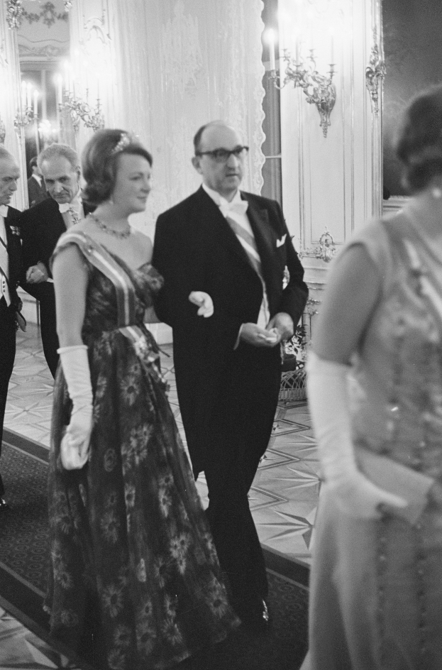 Ein Mann in einem schwarzen Blazer, weißem Hemd und schwarzen Hosen sowie eine Frau in einem eleganten Kleid gehen einen Flur entlang, umgeben von einer Gruppe von Menschen, die auf dem Boden stehen, mit ein paar an der Wand angebrachten Lichtern, die eine gem├╝tliche Atmosphäre schaffen.