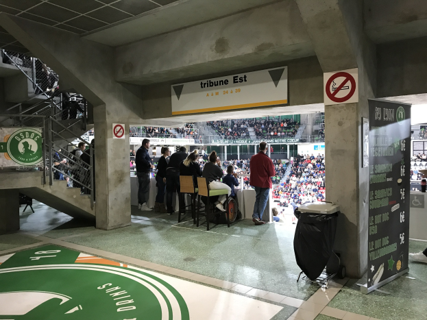 Eine Gruppe von Menschen steht vor einem sitzenden Publikum in einem Stadion, mit Schildern, die "Boston Celtics vs New York Jets", ein Müllcontainer, eine Tafel mit Text, eine Treppe mit Geländern und ein Dach mit Deckenleuchten anzeigen.