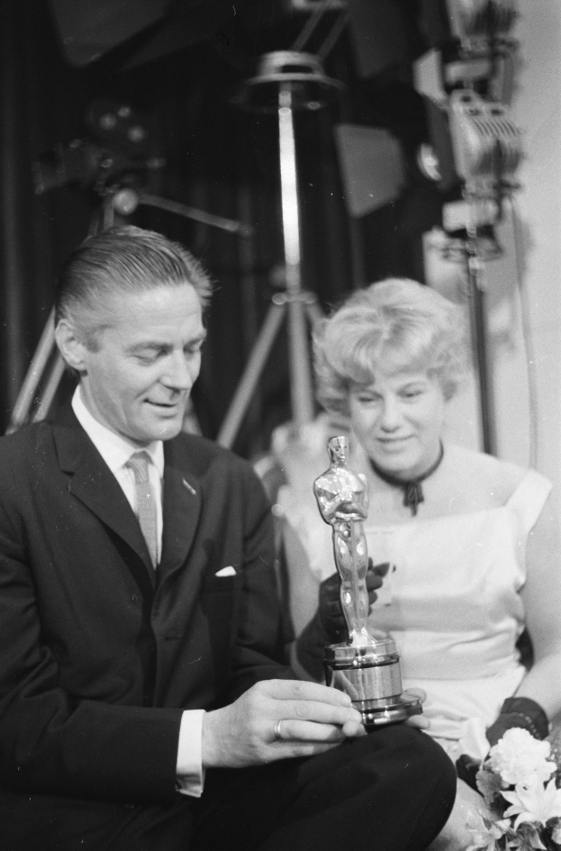 Ein Schwarz-Wei├č-Bild eines formell gekleideten Mannes mit einer Oscar-Statue und einer Frau mit einem Blumenstrau├č, die nebeneinander sitzen, mit beleuchtetem Hintergrund.