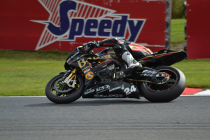 Eine Person mit Helm und Schuhwerk fährt ein Motorrad auf einer Rennstrecke, die von Gras umgeben ist, mit einer Tafel im Hintergrund, auf der Text angezeigt wird.