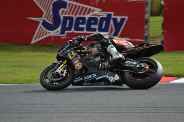 Eine Person mit Helm und Schuhwerk fährt ein Motorrad auf einer Rennstrecke, die von Gras umgeben ist, mit einer Tafel im Hintergrund, auf der Text angezeigt wird.