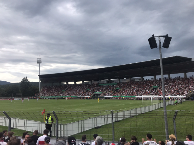 Große Zuschauermenge bei einem Fußballspiel in einem bewölkten, offenen Stadion mit beleuchtetem Spielfeld, umgeben von einem Zaun, Bäumen und Hügeln.