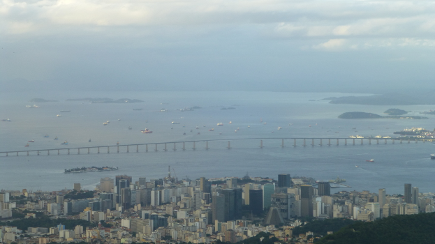 Luftaufnahme einer Stadtlandschaft mit Gebäuden, Bäumen, einem Gewässer mit Schiffen, einer Brücke und einem bewölkten Himmel.