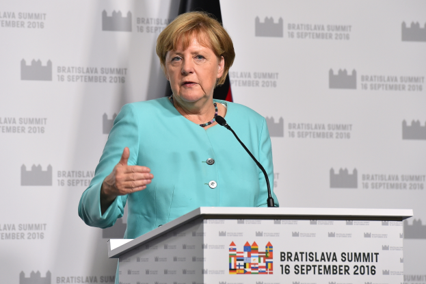 Bundeskanzlerin Angela Merkel hält eine Rede auf dem Bratislava-Gipfel, trägt ein blaues Kleid und steht vor einem Podium mit zwei Mikrofonen, mit einem Banner und einer Flagge im Hintergrund.
