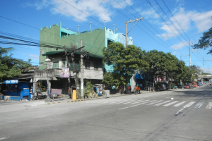 Eine Stadtstraße mit einem grünen Gebäude, fahrende Fahrzeuge, Fußgänger und Strommasten mit Drähten unter einem bewölkten Himmel.