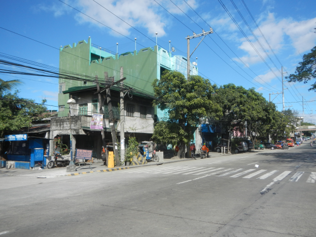 Eine Stadtstraße mit einem grünen Gebäude, fahrende Fahrzeuge, Fußgänger und Strommasten mit Drähten unter einem bewölkten Himmel.