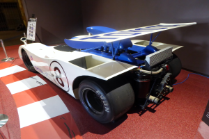 Ein weiß-blauer Rennwagen in einem Museum ausgestellt, umgeben von Absperrungen und Schautafeln mit Text, vor einer Wand, die auf das Goodwood Festival of Speed hindeutet.