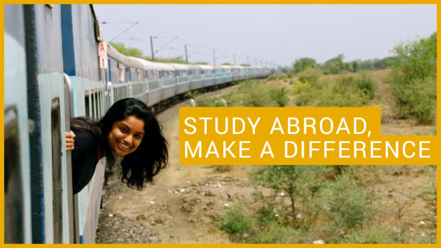 Eine Frau in einem schwarzen Kleid lächelt, während sie an einem Zug mit der Aufschrift "Study Abroad, Make a Difference" lehnt, vor einem Hintergrund aus Bäumen, Pfählen und einem klaren blauen Himmel.