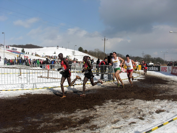 Eine Gruppe von Menschen, die bei einem Rennen auf einem schneebedeckten Feld laufen, mit einem Metallzaun, einer an einem Pfahl befestigten Schleife, einer Fahne mit Text, Zuschauern, Strommasten, Bäumen und einem bewölkten Himmel im Hintergrund.