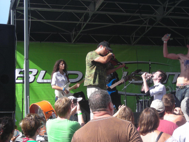 Eine Gruppe Musiker, die verschiedene Instrumente auf einer Bühne spielen, mit einem großen schwarzen Lautsprecher links daneben, während das Publikum vor einem Podium mit einem grünen Banner im Hintergrund steht.