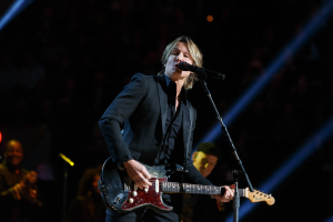 Keith Urban performs auf der Bühne mit einer Gitarre, einem Mikrofon und einer Band in einem schwach beleuchteten Veranstaltungsort.