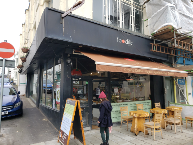 Eine Frau geht auf dem Gehweg an einem Gebäude mit Fenstern vorbei, das als Foodlic, ein veganes Restaurant in London, identifiziert wird, mit einem Schild, Topfpflanzen, Stühlen und Tischen, Fahrzeugen auf der Straße und einem bewölkten Himmel.