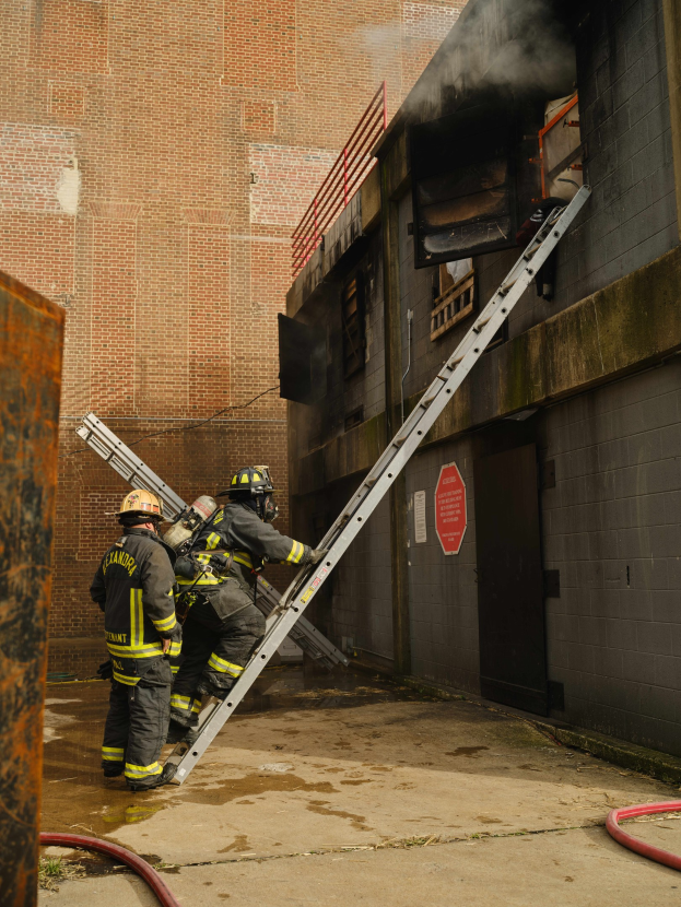 Feuerwehrleute mit Helmen, die eine Leiter hochsteigen und auf ein raucherfülltes Gebäude zusteuern, mit einem metallischen Objekt links und Rohren am Boden.