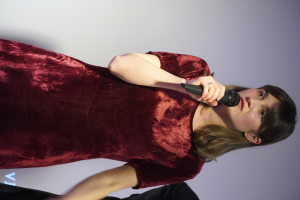 Eine Frau in einem roten Kleid steht selbstbewusst auf einer Bühne, hält ein Mikrofon und hat einen eleganten Bob-Haarschnitt und natürliches Make-up.