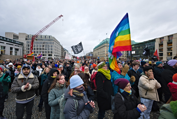 Eine große Gruppe von Menschen bei einer LGBTQ+-Rechtsdemo in Berlin, die Fahnen und Schilder schwenken, mit Gebäuden, einem Kran und einem bewölkten Himmel im Hintergrund.