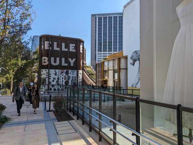 Menschen gehen auf einem Gehweg vor einem Gebäude mit einer Tafel, auf der "Elle Bully" steht, vorbei. Es gibt Bäume und Pflanzen auf beiden Seiten, eine Schaufensterpuppe auf der rechten Seite und andere Gebäude und einen klaren blauen Himmel im Hintergrund.