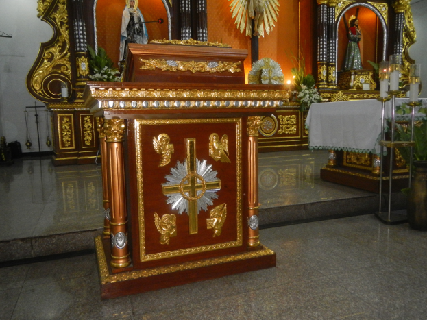 Ein Altar in einer Kirche mit einem Kreuz, Statuen, Blumensträußen, Kerzen mit Ständern, einem Tisch mit einer Decke und anderen Gegenständen, während die Wände mit Lichtern geschmückt sind.