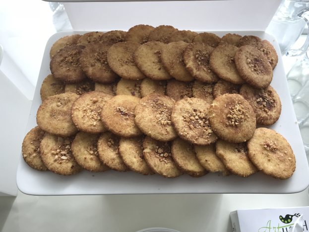 Ein weißer Teller voller Kekse auf einem Tisch, umgeben von Gläsern und anderen Gegenständen, mit einem Papier mit Text und Logo unten.