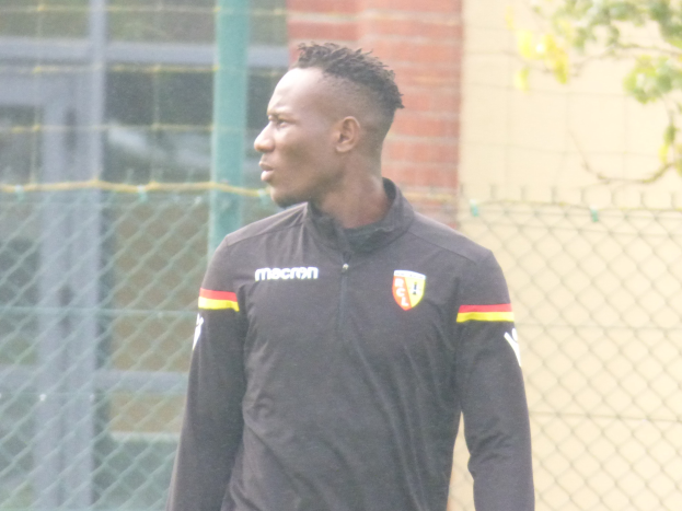 Ein Mann in einer schwarzen Jacke steht auf einem Fußballfeld, umgeben von einem Zaun und einem Baum, und sieht konzentriert während eines Trainings aus.