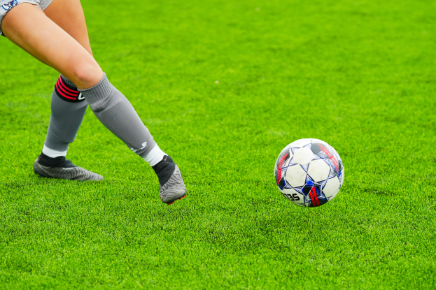 Eine Frau in Sportschuhen kickt einen Fussball auf einem saftig grünen Feld, was darauf hinweist, dass sie eine professionelle Fussballspielerin ist.