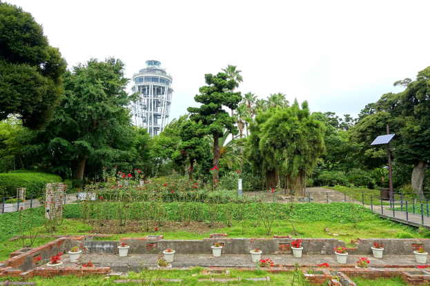 Ein Park mit einem Turm im Hintergrund, umgeben von grünem Gras, Pflanzen, Bäumen, Blumentöpfen, einer Straße mit Geländern und einem Pfahl mit einer Tafel unter einem sichtbaren Himmel.
