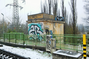 Ein Bahngeleis mit Graffiti an der Seite, umgeben von Gelöndern, Pfosten, Lichtern und Bäumen, mit Türmen und Drähten im Hintergrund und Schnee am Boden.