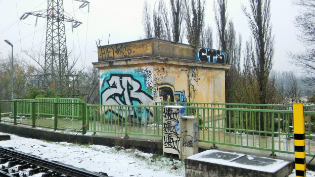 Ein Bahngeleis mit Graffiti an der Seite, umgeben von Gelöndern, Pfosten, Lichtern und Bäumen, mit Türmen und Drähten im Hintergrund und Schnee am Boden.