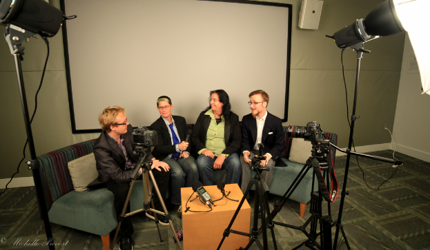 Eine Gruppe von drei Personen sitzt auf einem Sofa und gibt ein Interview, wobei eine Person in der Mitte ein Mikrofon hält, ein Aufnahmegerät auf dem Tisch in der Mitte steht, Kameras auf beiden Seiten und ein Projektionsschirm dahinter zu sehen ist.