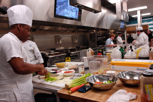 Eine Gruppe von Köchen in einer Küche, die Essen auf einem Schneidbrett zubereiten, mit einem Koch im Vordergrund in einem weißen Kleid und einer Mütze, der ein Messer hält und von verschiedenen Utensilien, Schüsseln, Gemüse, einem Handy, Flaschen und anderen Gegenständen umgeben ist, mit einem Fernseher und Deckenleuchten im Hintergrund.