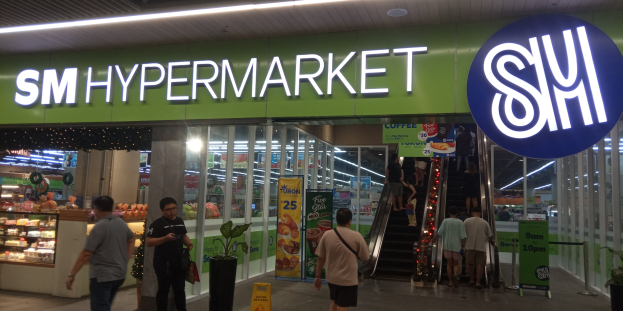 Eine Gruppe von Menschen vor einem hell erleuchteten "SM Hypermarket"-Laden mit sichtbarer Beschilderung, Texttafeln und Topfpflanzen.