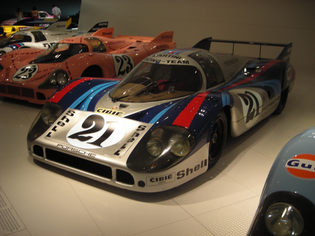 Ein Porsche 917K Rennsportwagen im Museum ausgestellt, umgeben von anderen Sportwagen und Menschen, mit einer Wand und informativem Text im Hintergrund.