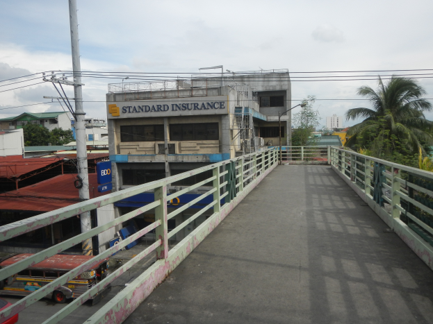 Eine Brücke mit Geländern, Fahrzeuge auf der Straße, Gebäude, Bäume, Strommasten mit Drähten und ein bewölkter Himmel im Hintergrund, mit einem Gebäude, auf dem ein Schild mit der Aufschrift "Standard Insurance" steht.