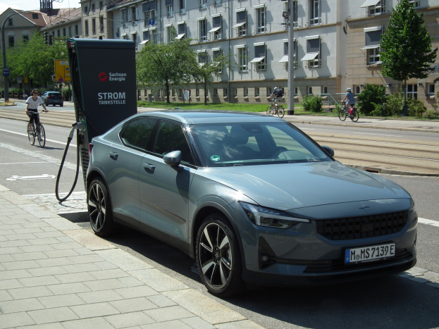 Elektroauto neben einer Ladesäule an einer Straße mit Radfahrern auf dem Gehweg, Bäumen, Gebäuden, Laternen und einem bewölkten Himmel im Hintergrund.