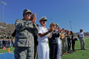 Eine Gruppe von Menschen auf einem Fußballfeld klatschen, mit Zuschauern in den Rängen, Flutlichtmasten und einem klaren blauen Himmel im Hintergrund.