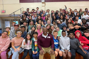 Gruppe von Schülern, die für ein Foto in einer Turnhalle posieren, einige sitzen auf der Treppe und andere stehen, eine Tür und ein Ausgangsschild links sichtbar