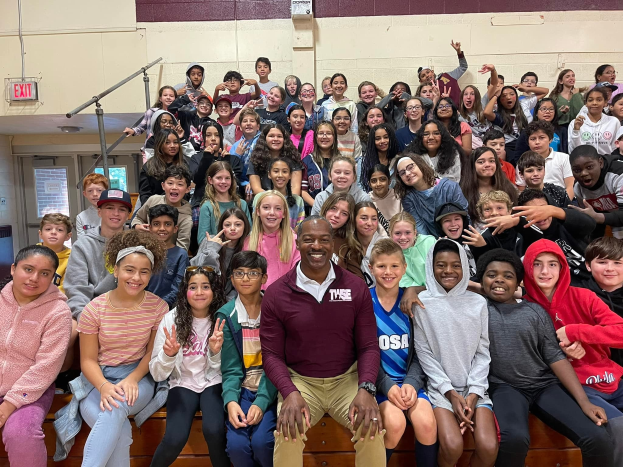 Gruppe von Schülern, die für ein Foto in einer Turnhalle posieren, einige sitzen auf der Treppe und andere stehen, eine Tür und ein Ausgangsschild links sichtbar