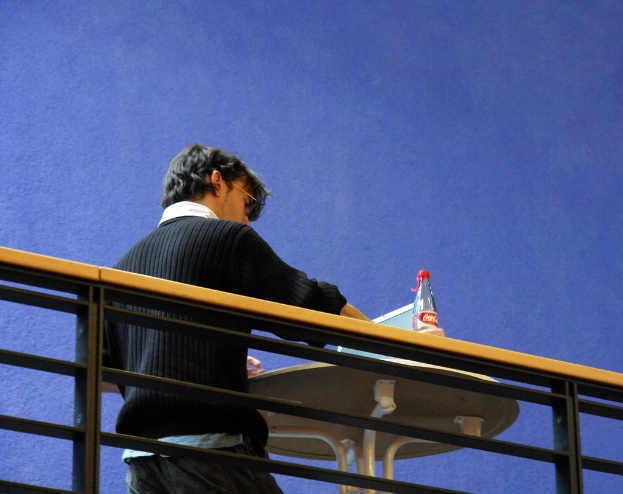 Eine Person in einem schwarzen Hoodie steht an einem Tisch mit einem Laptop und einer Flasche, mit einem blauen Gemälde an der Wand im Hintergrund und einem Geländer im Vordergrund.