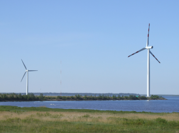 Drei Windkraftanlagen in einer grünen Wiese in der Nähe von Wasser, mit Bäumen und Türmen im Hintergrund unter einem klaren blauen Himmel.