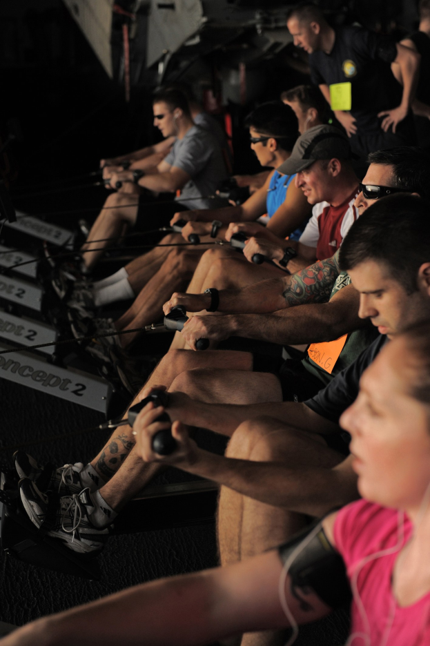 Eine Gruppe von Menschen, die an Rudermaschinen in einem Fitnessstudio sitzen, mit anderen im Hintergrund, die alle sportliche Schuhe tragen.