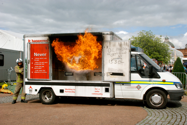 Ein brennender Transporter mit Schrift darauf im Vordergrund, mit einem Mann dahinter und einem Weg, der zu Gebäuden, Menschen, Pflanzen, einem Baum und einem bewölkten Himmel führt.