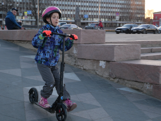 Ein junger Junge fährt mit einem Roller eine Straße entlang, trägt einen Helm und Handschuhe und hat verschiedene urbane Elemente und einen klaren blauen Himmel im Hintergrund.