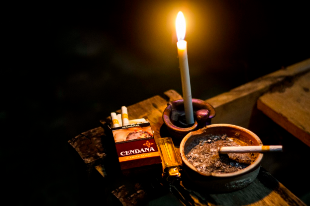 Eine Kerze, eine Packung Zigaretten, eine Schachtel, ein Feuerzeug und ein Behälter sind auf einer Oberfläche angeordnet.