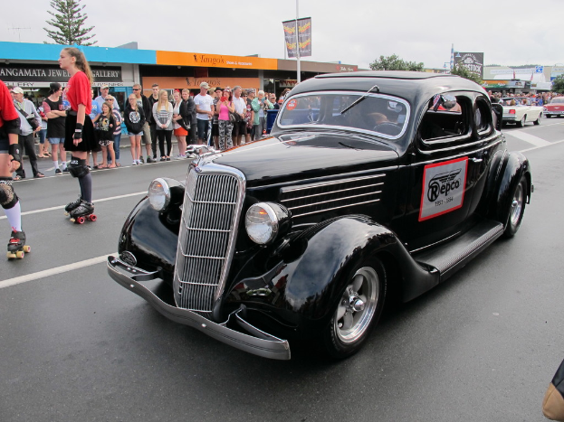 Eine Straßenszene mit ein paar Autos, Menschen, einige mit Mützen, Gebäuden, Bäumen, einem Werbeplakat, ein paar Individuen mit Rollschuhen, einem bewölkten Himmel und einem Banner an einem Pfahl.