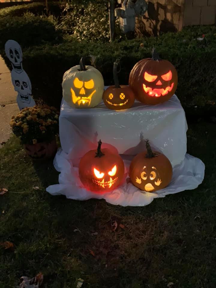 Eine Gruppe von geschnitzten Kürbissen auf einem weißen tuchbedeckten Tisch umgeben von Gras und trockenen Blättern, mit Pflanzen, Bäumen und einer Wand im Hintergrund.