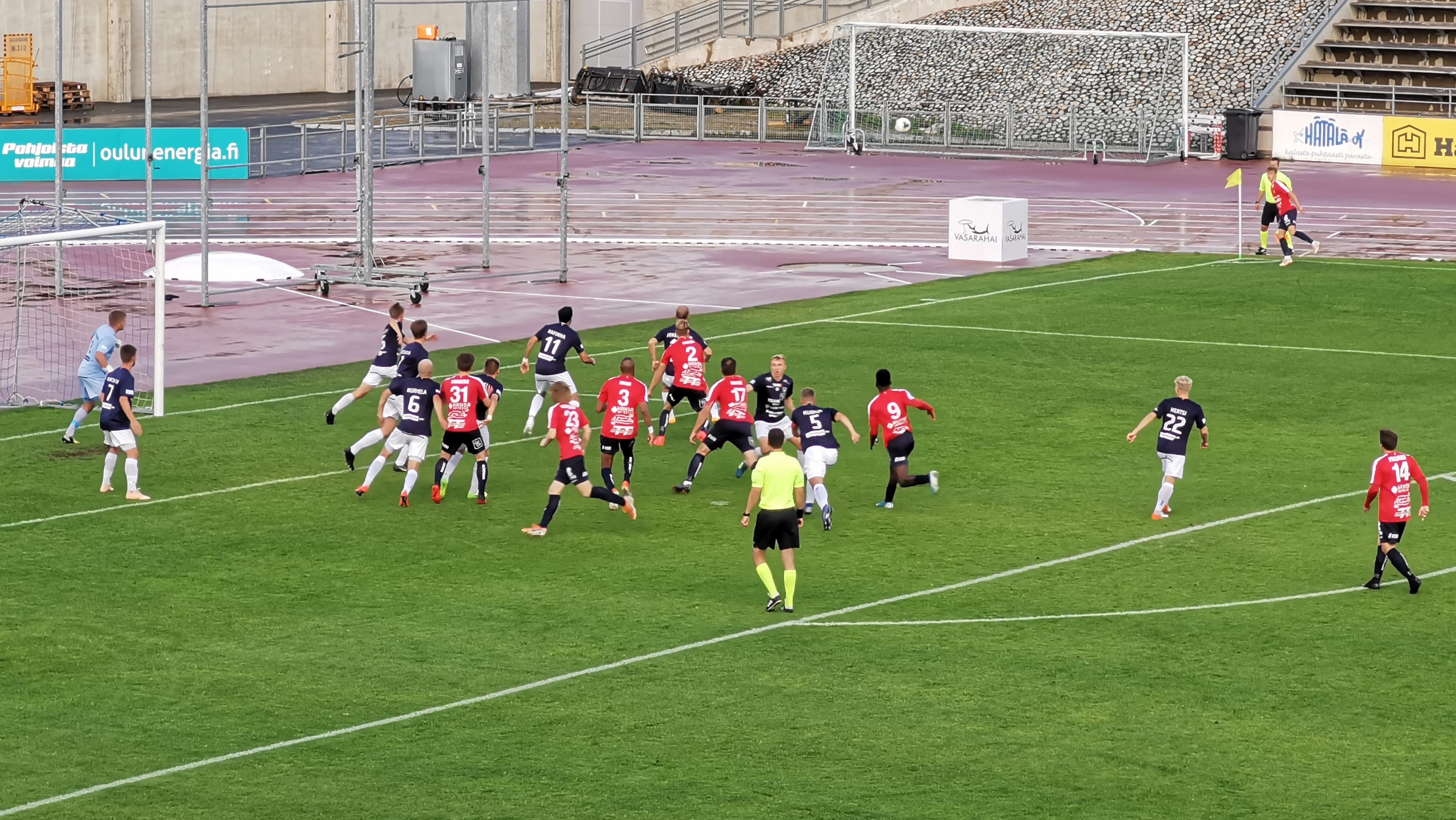 Gruppe von Menschen, die Fußball auf einem Feld mit Zuschauern in einem Stadion spielen, umgeben von Geländern, Pfosten und Beschilderung.