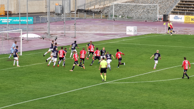 Gruppe von Menschen, die Fußball auf einem Feld mit Zuschauern in einem Stadion spielen, umgeben von Geländern, Pfosten und Beschilderung.