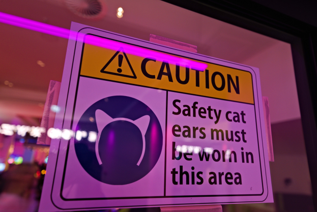Ein Warnschild in einem Schaufenster trägt die Aufschrift "Vorsicht: Sicherheitskatzenohren müssen in diesem Bereich getragen werden." vor einem unscharfen Hintergrund.