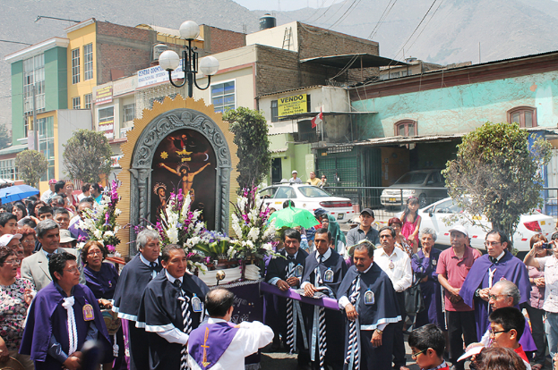 Menschen auf einer Straße mit einem Kruzifix und Blumensträußen im Vordergrund, Autos in der Mitte und Gebäude, Bäume und Straßenlaternen im Hintergrund.
