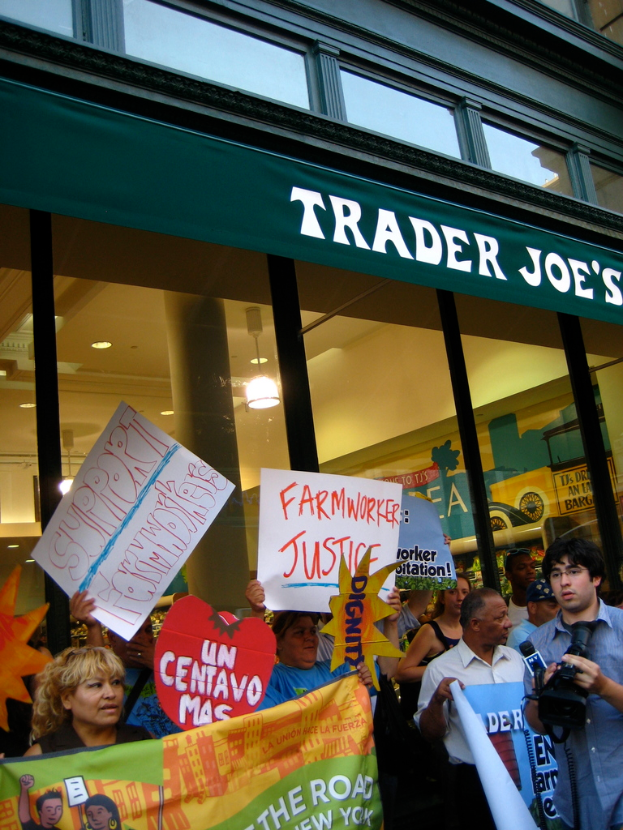 Gruppe von Menschen vor einem Geschäft mit Schildern und Plakaten, im Hintergrund ist eine Säule und eine Dachbeleuchtung zu sehen.